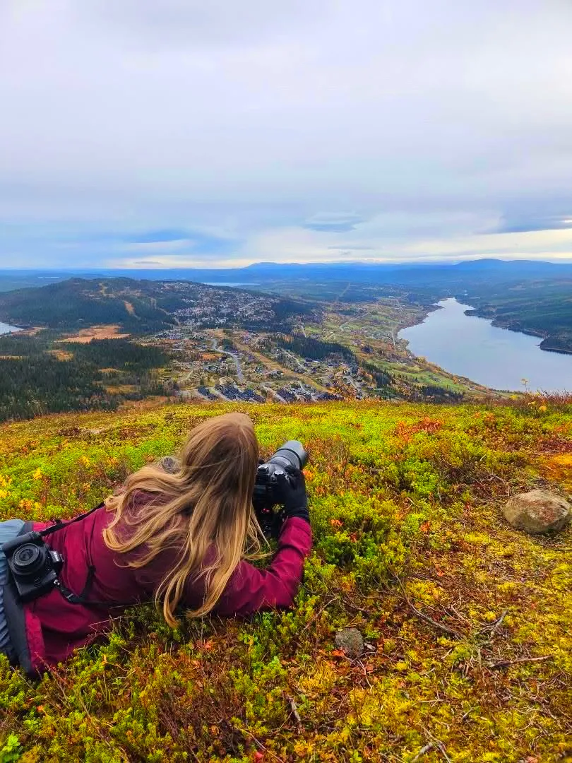 Josefin ligger i fjällgräset med kameran och fotograferar utsikten över dalen